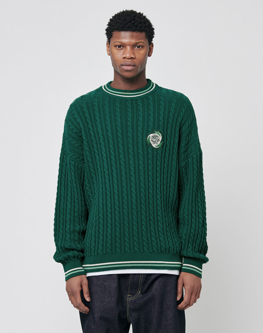 A young man wears a LIVE FAST DIE YOUNG Knitted Sweater in oversize fit, dark green with cable knit, white and green striped cuffs and collar, and an embroidered chest patch, standing against a plain light gray background.