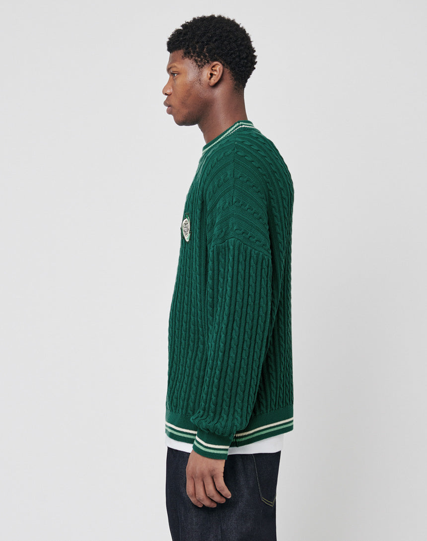 A young man stands in profile against a plain background, wearing a LIVE FAST DIE YOUNG Knitted Sweater in dark green with white trim, oversize fit, paired with dark jeans.