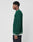 A young man stands in profile against a plain background, wearing a LIVE FAST DIE YOUNG Knitted Sweater in dark green with white trim, oversize fit, paired with dark jeans.