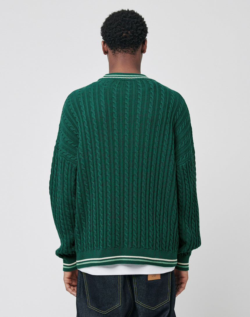A person wearing a dark green LIVE FAST DIE YOUNG Knitted Sweater with cable pattern and white trim, in an oversize fit, stands facing away from the camera. The sweater is paired with dark jeans against a light gray background.