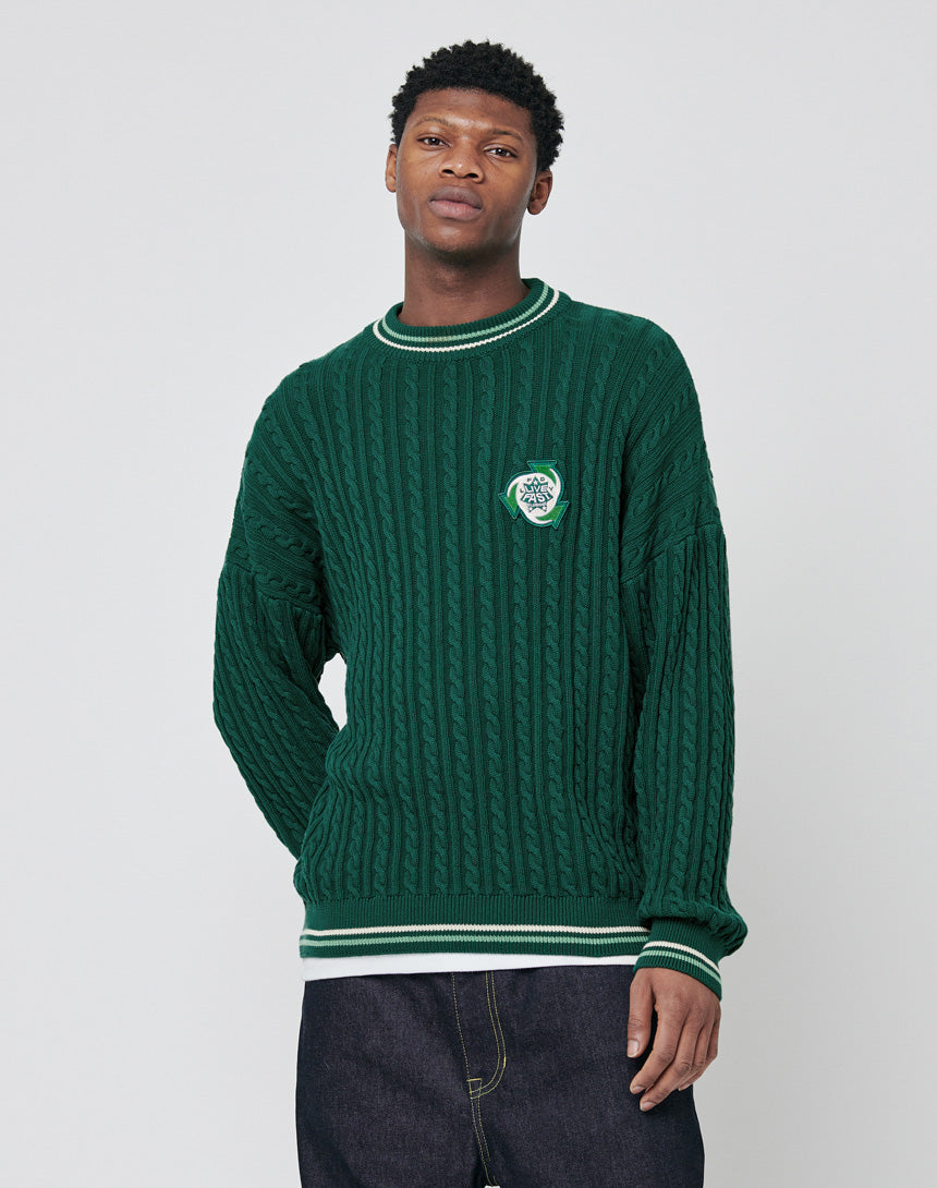 A young man in an oversize fit, dark green LIVE FAST DIE YOUNG Knitted Sweater with white trim and a small chest patch stands against a light gray background. He has short curly hair and wears dark jeans.