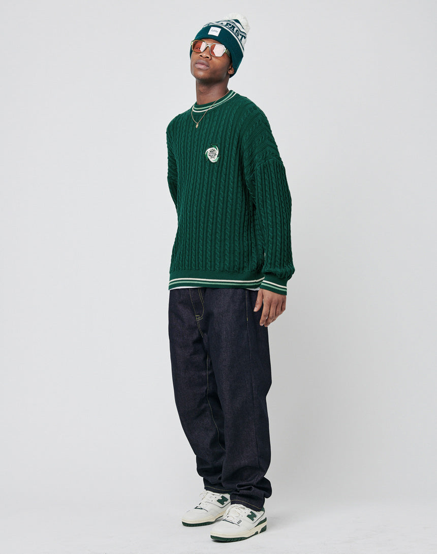 A person poses against a plain background wearing a green LIVE FAST DIE YOUNG Knitted Sweater, dark baggy jeans, white sneakers, a green and white beanie, sunglasses, and layered gold necklaces.