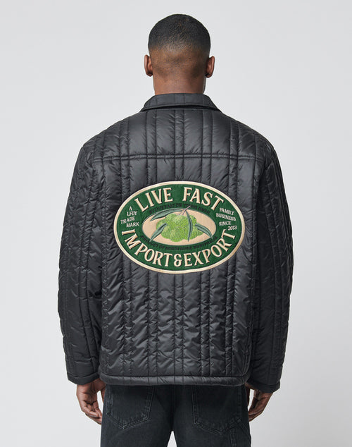 A man wears the LIVE FAST DIE YOUNG Import & Export Jacket—black, oversize fit, lightly padded, with a large green and beige patch on the back reading LIVE FAST IMPORT & EXPORT and featuring smaller text plus a green car graphic.
