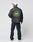 A man in the LIVE FAST DIE YOUNG Import & Export Jacket, featuring a bold oval patch with a green mountain graphic, pairs it with black jeans and a white cap as he stands facing away.