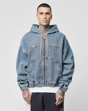 A person with short hair and tattoos wears the LIVE FAST DIE YOUNG Denim Hooded Zip—a loose-fit blue denim hoodie with a zip, hood, and kangaroo pocket—against a plain light background.