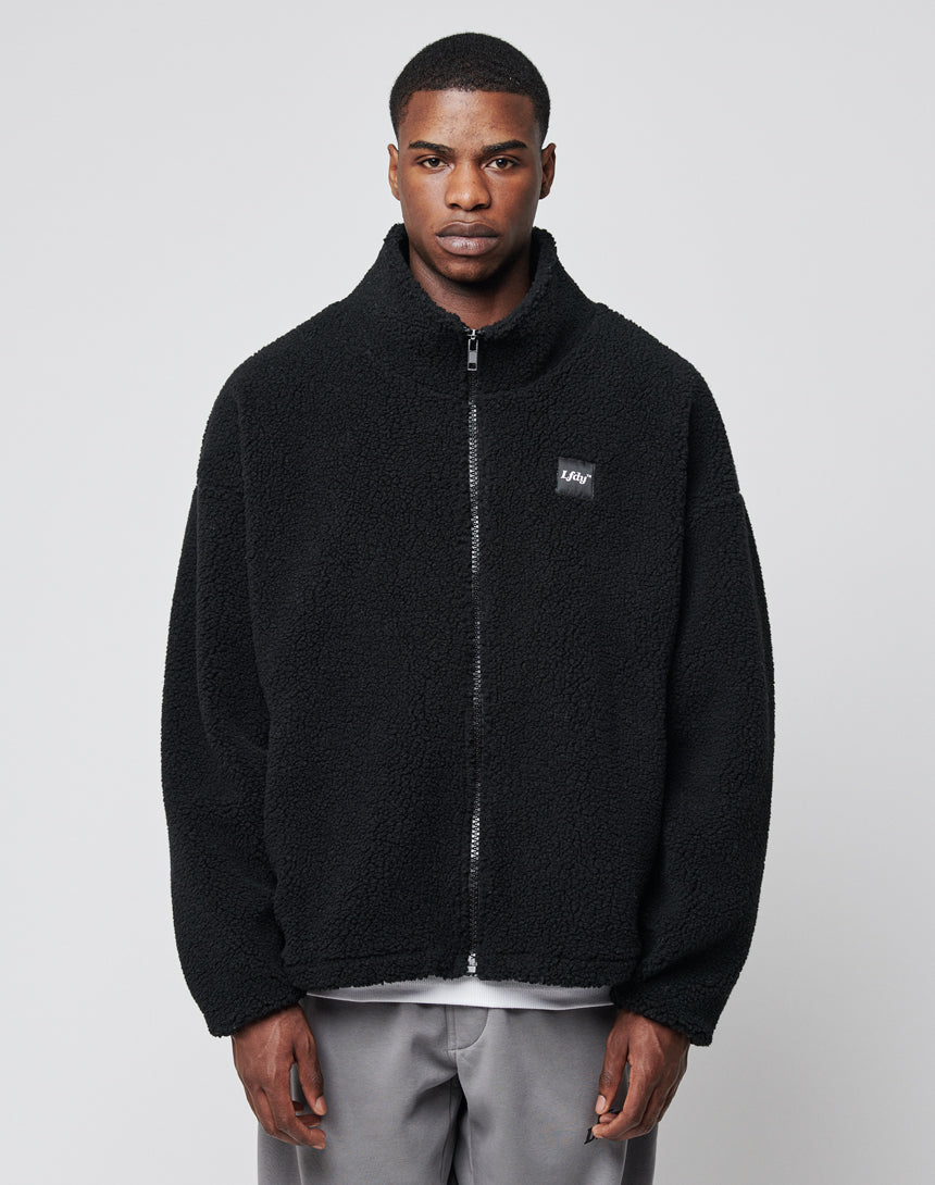 A man faces forward, wearing the LFDY Oversize Teddy Jacket in black with a wide collar and small chest logo, paired with light gray pants, set against a plain light gray background.