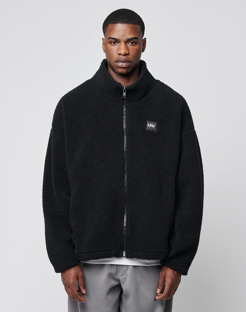 A man faces forward, wearing the LFDY Oversize Teddy Jacket in black with a wide collar and small chest logo, paired with light gray pants, set against a plain light gray background.