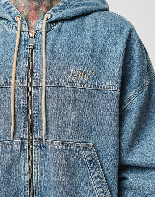 Close-up of a person in the LIVE FAST DIE YOUNG Denim Hooded Zip, a blue denim zip-up with beige drawstrings, Lfdyl™ chest embroidery, kangaroo-tasche, oversize fit, and 100% Baumwolle. Colorful neck tattoos are visible.