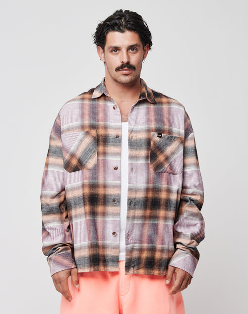A dark-haired man with a mustache wears the LIVE FAST DIE YOUNG Loose Summer Flannel—an oversize pink, brown, and gray plaid shirt—over a white tee and light pink pants, standing against a plain light background.
