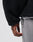 A close-up of a hand resting by the side, wearing the LFDY Oversize Teddy Jacket in black with adjustable drawstring and weiter Kragen, paired with light gray pants against a plain, unfocused background.