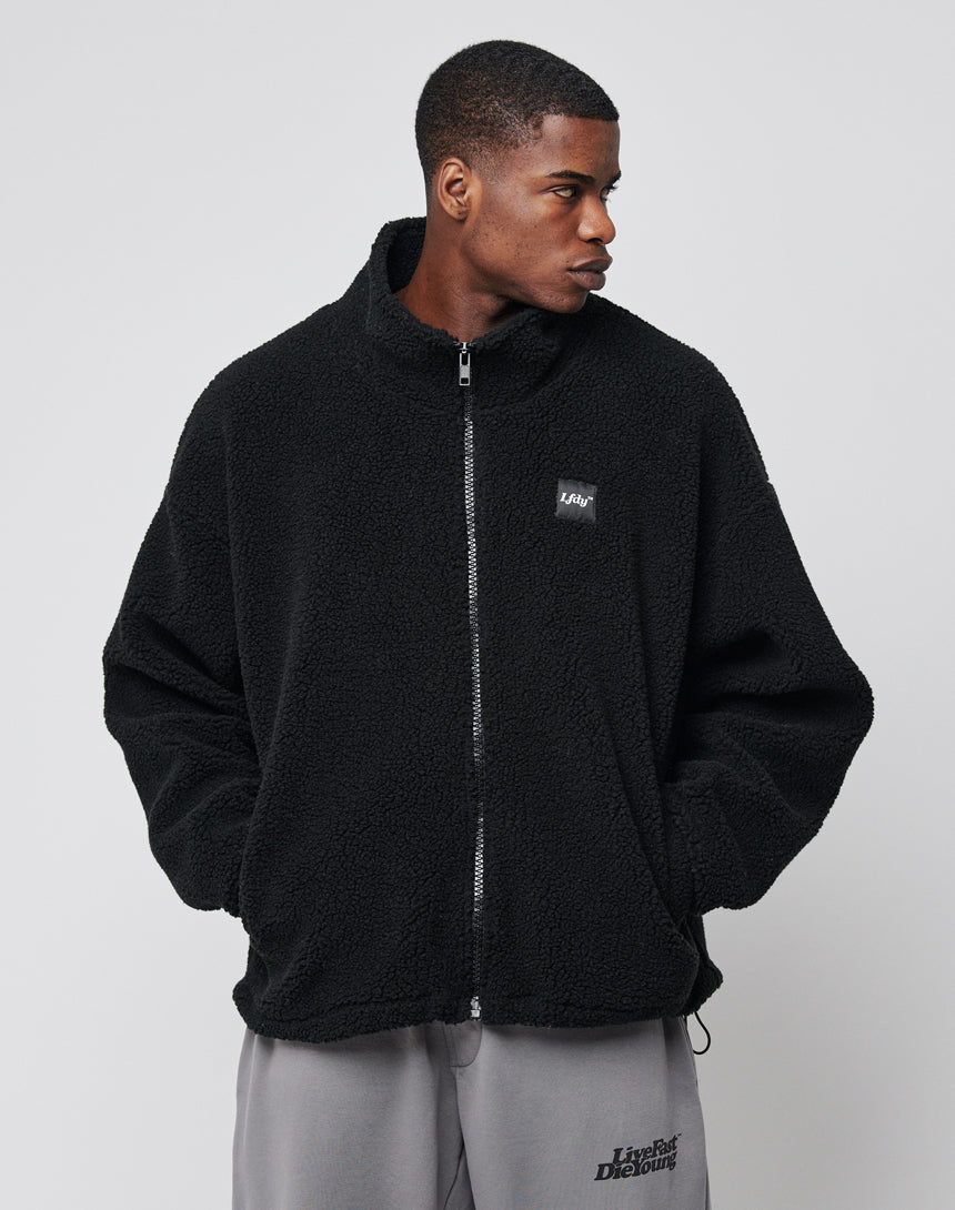 A man stands with his hands in his pockets, wearing the LFDY Oversize Teddy Jacket in black with a small chest logo and gray sweatpants, looking to the side against a plain light gray background.