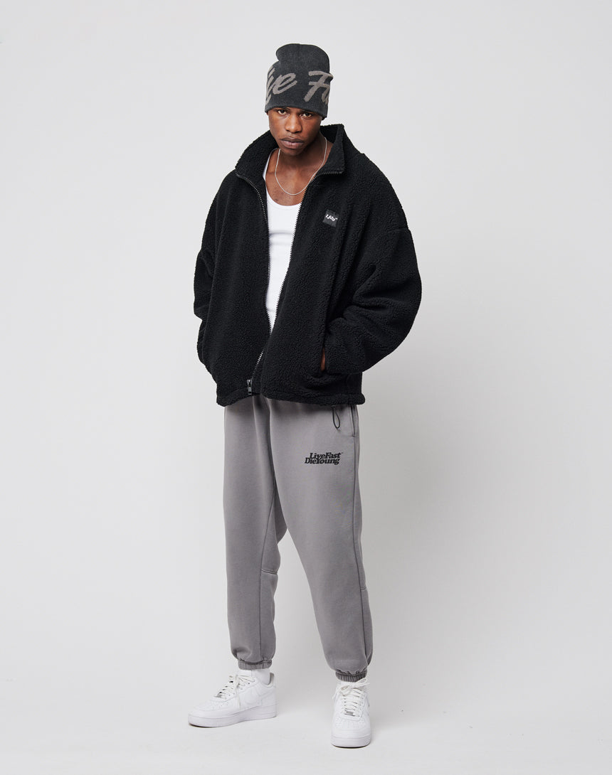 A man stands against a plain background wearing a gray beanie, white tank top, gray sweatpants, white sneakers, and an LFDY Oversize Teddy Jacket with his hands in the jacket pockets and a serious expression.