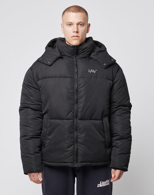 A person stands facing forward in a LIVE FAST DIE YOUNG Basic Winter Jacket with an oversized fit, detachable hood, and Idu on the chest, paired with dark pants featuring white text on the left leg. The background is plain light gray.