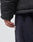 A close-up of a person’s hand by their side, wearing the LIVE FAST DIE YOUNG Basic Winter Jacket in black oversize fit and dark blue sweatpants with partially visible white text. The background is simple and bright.