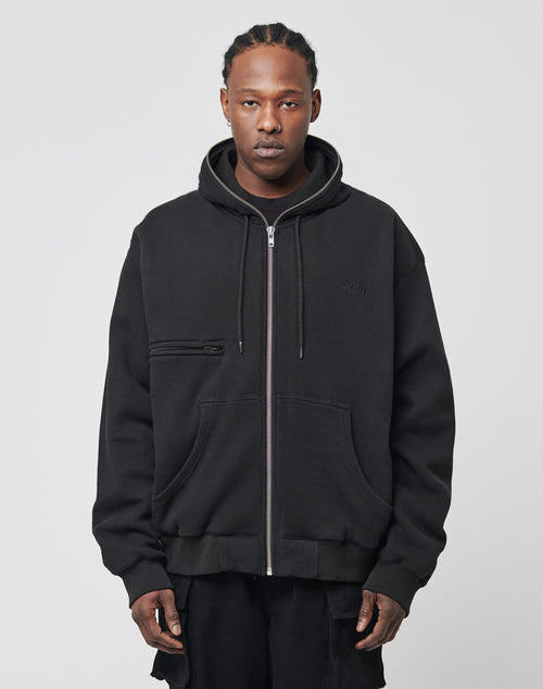 A person faces forward in a black Waffle Hooded Zip by LIVE FAST DIE YOUNG, featuring an oversize fit, chest pocket, and subtle Logo-Stick, layered over a black shirt against a light plain background.