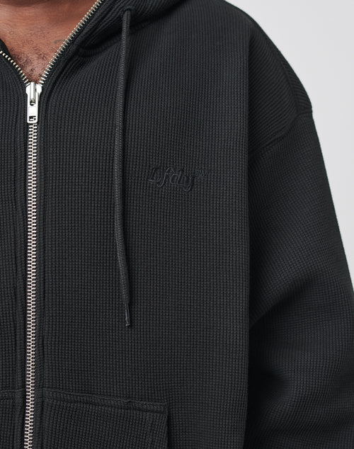 A close-up shows part of the chest and shoulder of a person wearing the LIVE FAST DIE YOUNG Waffle Hooded Zip, a black oversize fit hoodie made from waffle-textured material with drawstrings, a front pocket, and embossed logo text on the chest.