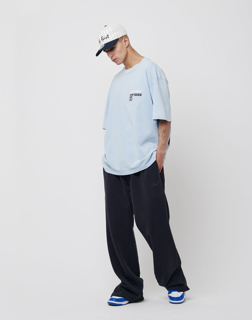 A person wearing a light blue oversized T-shirt, LFDY Wide Jogger 2.0 in black, blue-and-white sneakers, and a white cap stands against a plain white background, looking down with hands in their pockets.