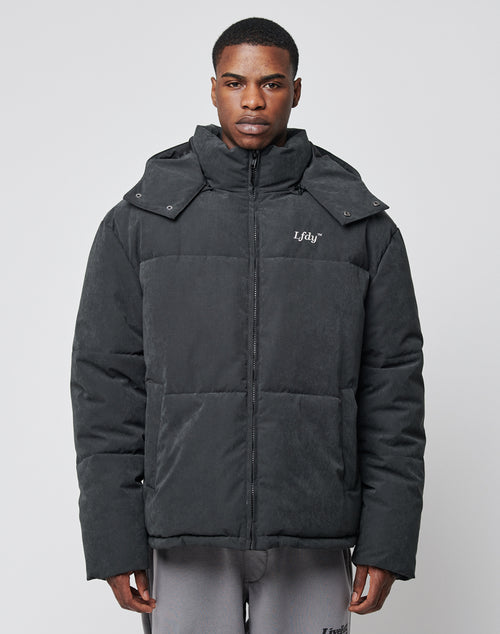 A man stands facing forward in a dark grey, oversize fit LIVE FAST DIE YOUNG Basic Winter Jacket with high collar and detachable hood, paired with light grey sweatpants against a plain light background.