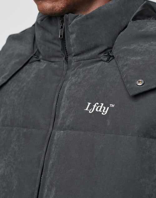 Close-up of a person wearing the LIVE FAST DIE YOUNG Basic Winter Jacket, a dark, wind- and weatherproof padded jacket with a high collar, visible zipper, snap buttons, and the white LFDY™ logo on the chest.
