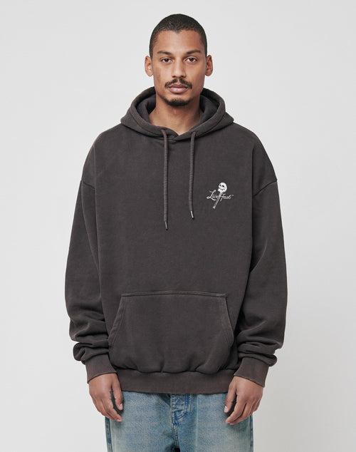 A man stands facing forward against a plain light grey background, wearing the LIVE FAST DIE YOUNG Live Fast Skull Hooded in dark grey with a subtle white logo and vintage effect, paired with light blue jeans for a relaxed look.