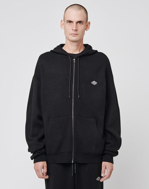 A man with a shaved head wears the LIVE FAST DIE YOUNG Knit Hooded Zip, an oversize black hoodie with a small chest logo patch and hood, standing against a plain white background.
