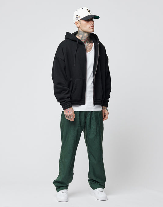 A man in a black zip-up hoodie over a white tank top, LIVE FAST DIE YOUNG Nylon Jogger pants with Breiter Elastikbund, white sneakers, and a white cap stands against a light gray background, showing tattoos on his neck and hands.
