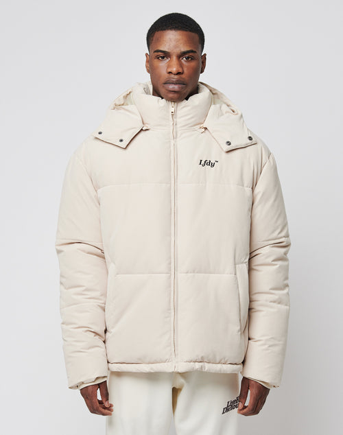 A man stands facing forward in a light beige LFDY Basic Winter Jacket with hood and chest logo, paired with matching pants. The oversize wind- and weatherproof jacket is shown against a plain white background.