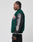 A man with braided hair is wearing the LIVE FAST DIE YOUNG Life College Jacket (LF2210-13153), a dark green varsity style with black leather sleeves, terry patches, a brain chest patch, and a skeletal hand and forearm sleeve patch. He stands sideways.