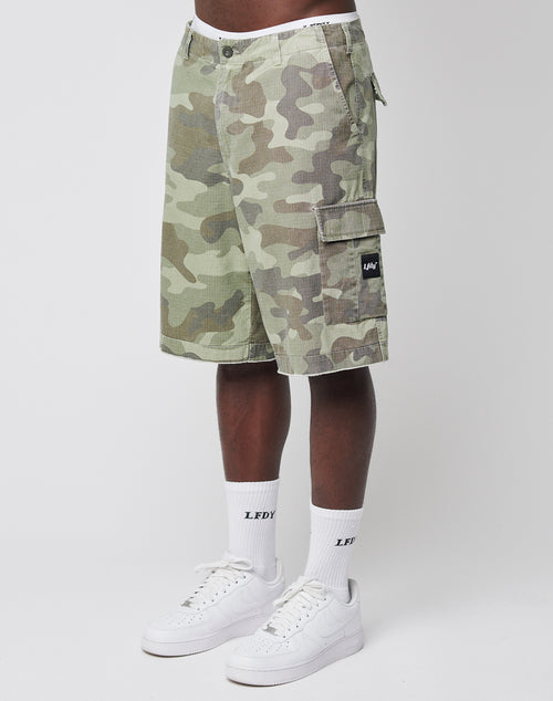A person wearing LIVE FAST DIE YOUNG Army Shorts, white crew socks with LEDY written on them, and white Nike sneakers stands against a plain white background.
