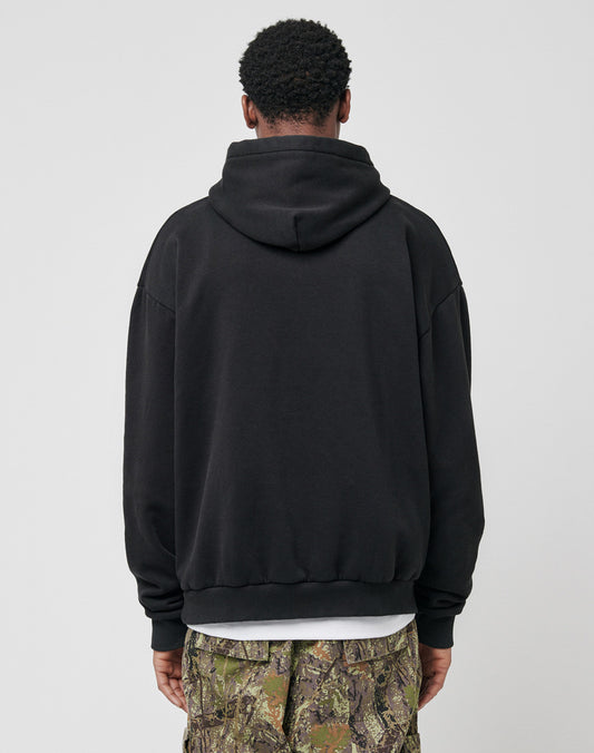 A person faces away from the camera, wearing the LIVE FAST DIE YOUNG Live Fast Rhinestone Hooded—an oversized black hoodie with a rhinestone logo—and camouflage pants, set against a plain light gray background.
