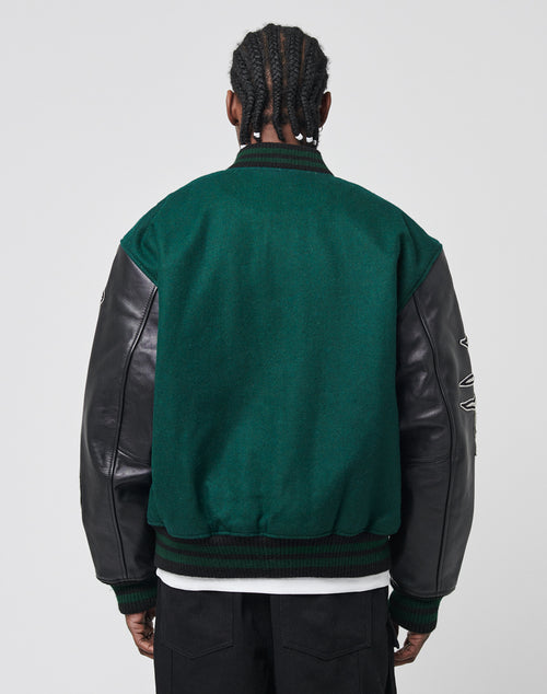 A person with braided hair is seen from the back, wearing the LIVE FAST DIE YOUNG Life College Jacket, featuring green fabric, black leather sleeves, logo snap buttons, and paired with black pants against a plain white background.