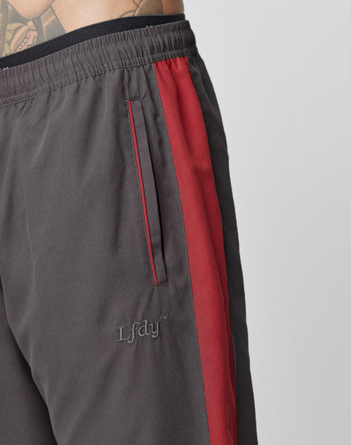 Close-up of a person wearing gray LFDY Trackpants by LIVE FAST DIE YOUNG, featuring a red side stripe and embroidered logo near the pocket; tattooed skin appears above the waistband.