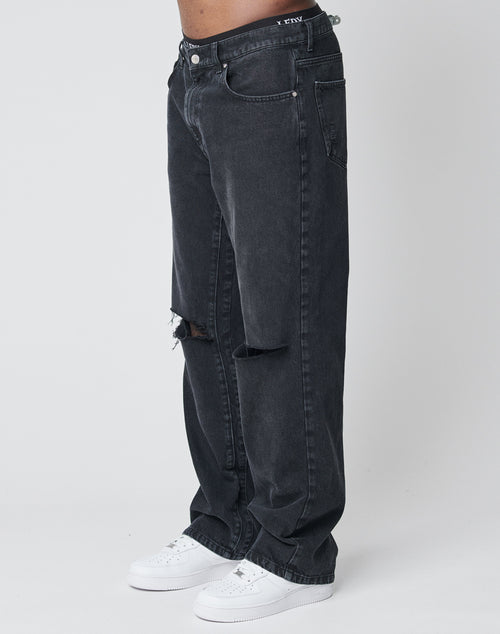 A person wears LIVE FAST DIE YOUNG Oversize Ripped Denim—loose-fitting black jeans with ripped knees—paired with white sneakers, standing against a plain white background.