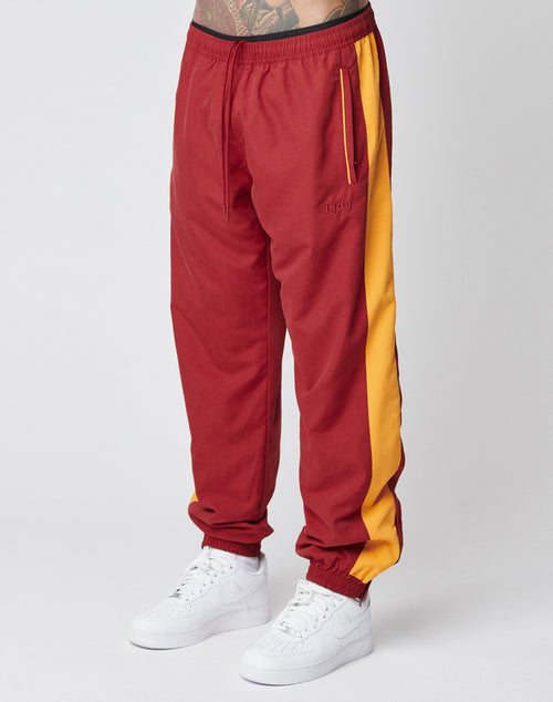 A person wears LIVE FAST DIE YOUNG LFDY Trackpants—regular fit, red with yellow side stripe, zipped Reißverschluss pockets, elastic waistband—made from polyester. Theyre paired with white sneakers against a plain light gray background.