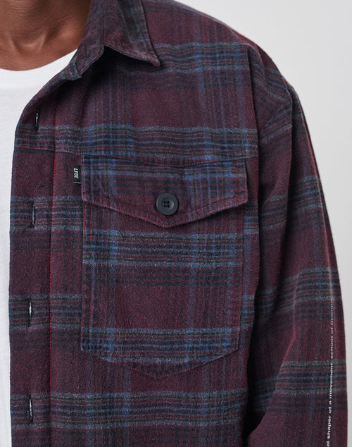 Close-up of a person wearing the LIVE FAST DIE YOUNG Padded Flannel Jacket in maroon and navy plaid, featuring a black buttoned chest pocket over a white t-shirt. Only the jacket and part of the neck and chin are visible.