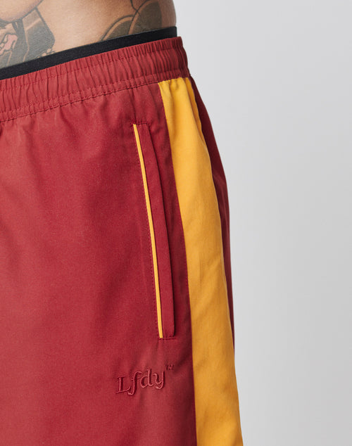 A person sports LIVE FAST DIE YOUNGs LFDY Trackpants: red polyester with yellow side stripes, LFDY embroidery by the pocket, and a comfy elastic waistband. Tattoos are partly visible on their torso.