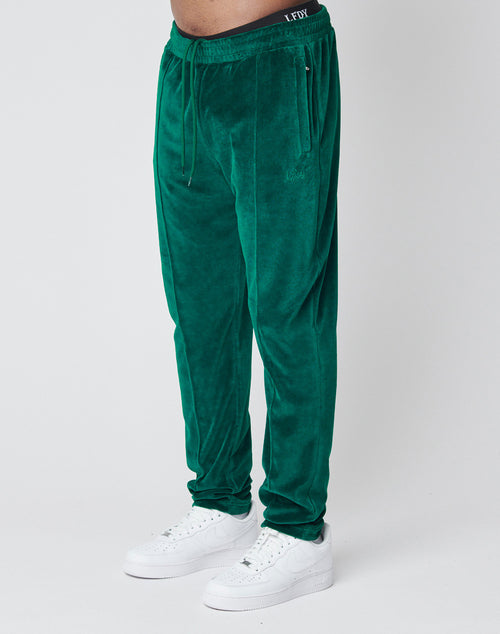 A person wearing LIVE FAST DIE YOUNG Velvet Tracksuit Pants with an elastic waistband, white sneakers, and black logo-stitched underwear stands against a plain white background.