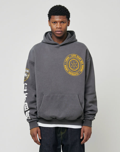 A young man wears the LIVE FAST DIE YOUNG LFDY Department Hooded in dark gray, featuring a yellow circular front print, a sleeve patch, and white text down the right sleeve, standing against a plain light background.