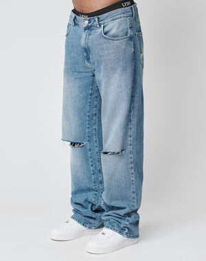 A person wears LIVE FAST DIE YOUNG Oversize Ripped Denim in light blue with knee slits, white sneakers, and a visible black waistband with white text. The plain white background highlights the relaxed vibe.