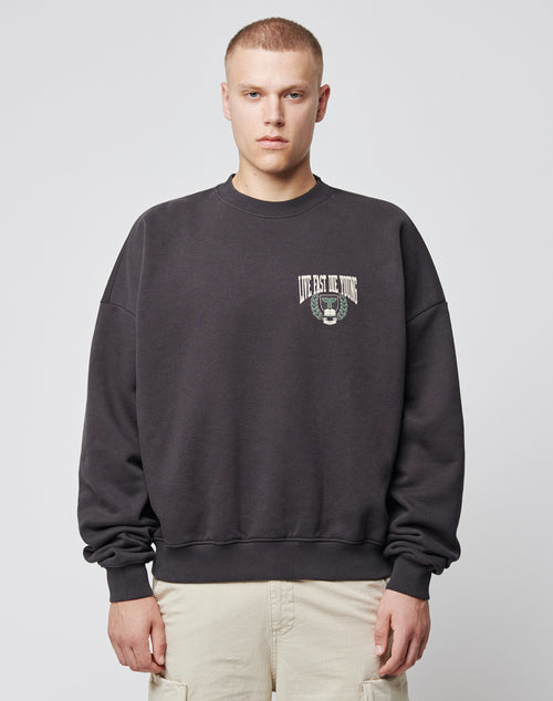 A person wears the LIVE FAST DIE YOUNG Live Fast Die Young Chest Sweater, a black oversized piece featuring a vintage LIVE FAST DIE YOUNG chest print and left-chest graphic, standing against a light gray background.