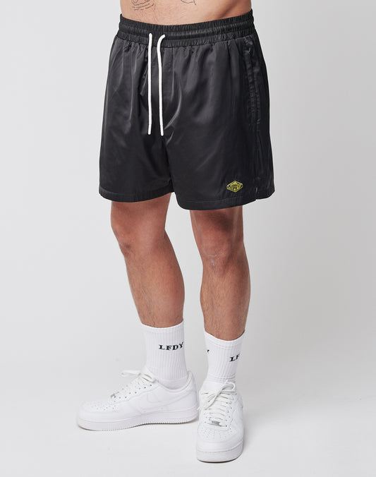 A person models LIVE FAST DIE YOUNG Basic Satin Shorts in black with a small yellow and green logo, paired with white crew socks featuring LEDY text and white sneakers. Only the lower half is visible against a plain white background.