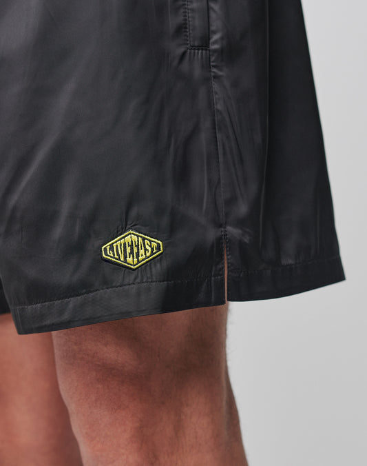 A close-up shows a person wearing LIVE FAST DIE YOUNG’s Basic Satin Shorts in black with a regular fit, elastic waistband, and a small yellow-green diamond-shaped LIVEFAST patch near the hem against a plain light background.