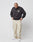 A man stands against a plain white background, looking down. He wears the LIVE FAST DIE YOUNG Live Fast Die Young Chest Sweater (black, oversized with chest print), beige cargo pants with side pockets, and white sneakers.