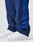 A close-up shows a person wearing LIVE FAST DIE YOUNG LFDY Trackpants—blue with a lighter side stripe and an elastic waistband. Made from polyester in a regular fit, the background is plain and light-colored.