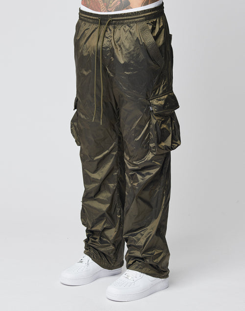 A person wearing LIVE FAST DIE YOUNG Parachute Cargo shiny olive green nylon pants with multiple cargo pockets and white sneakers stands against a plain light background; only their lower body is visible.