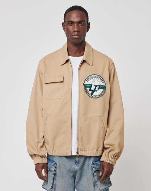 A man faces forward in a LIVE FAST DIE YOUNG Live Fast Twill Jacket, Regular Fit beige zip-up with Hemdkragen and a green-white circular patch on the left chest, over a white shirt and blue jeans with large side pockets, against a plain white background.