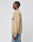 A person stands in profile against a plain background, wearing the LIVE FAST DIE YOUNG Live Fast Twill Jacket in tan with a green and white Stickapplikation logo on the chest, paired with light blue Regular Fit jeans with visible seams.