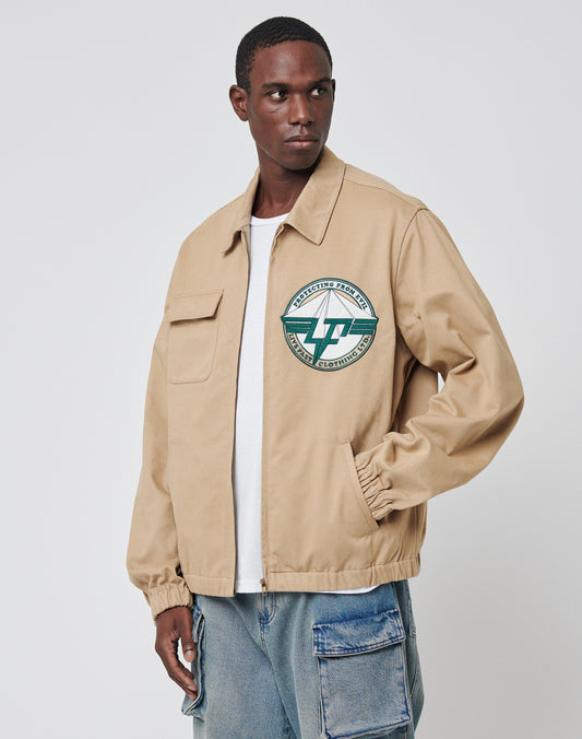 A man in a beige Live Fast Twill Jacket by LIVE FAST DIE YOUNG, featuring a Hemdkragen and chest emblem, paired with a white T-shirt and light blue patchwork jeans, stands against a plain white background.