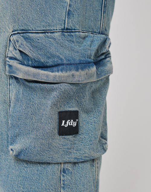 Close-up of the Wide Denim Cargo pocket by LIVE FAST DIE YOUNG in light blue, with a black patch label featuring white Lfdv text on the flap, subtle vintage effects, and an oversized fit.