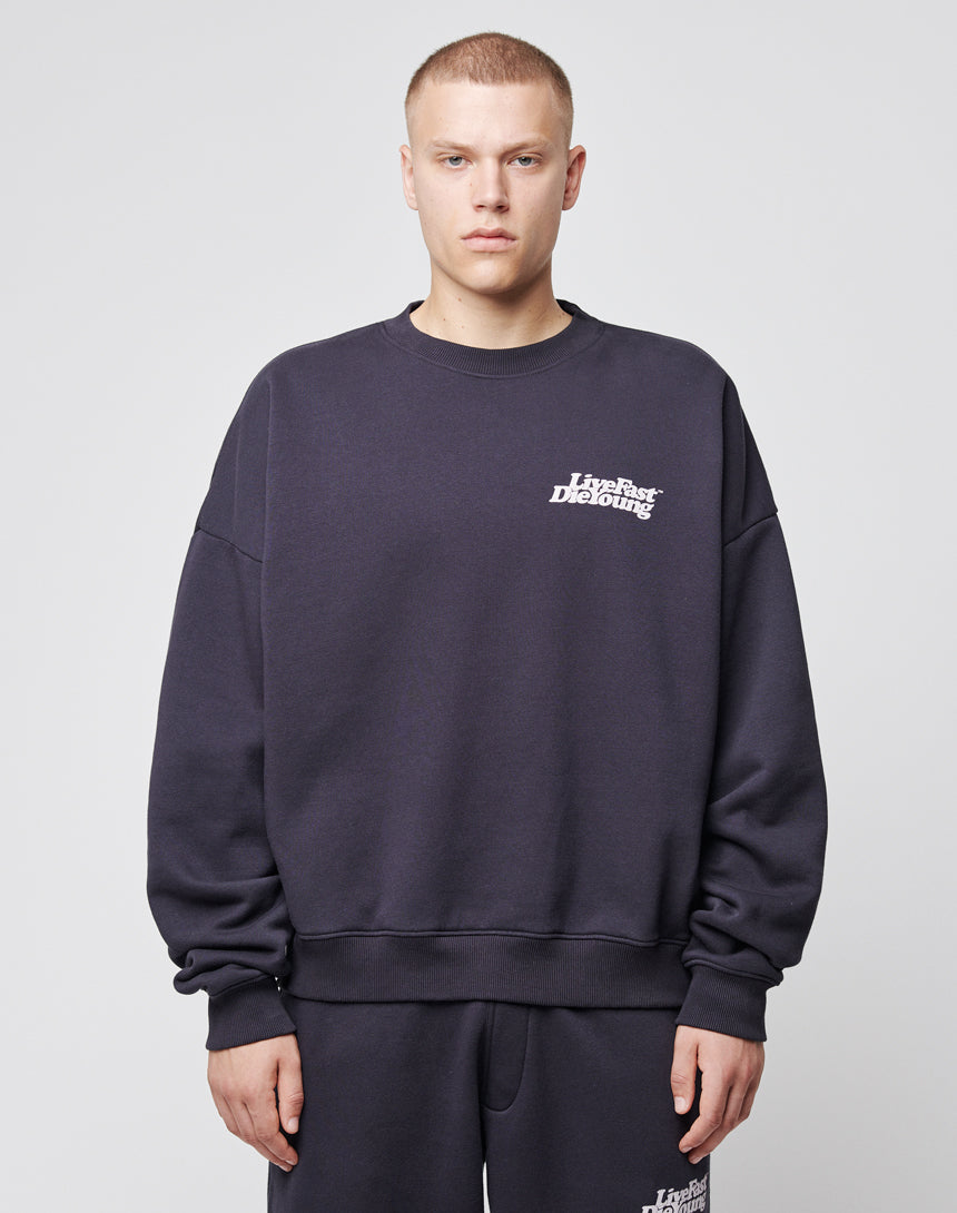 A person with short hair wears a loose-fitting Basic Chest Sweater by LIVE FAST DIE YOUNG and matching pants. The sweater features a small white chestprint logo on the left side. The background is plain and light-colored.